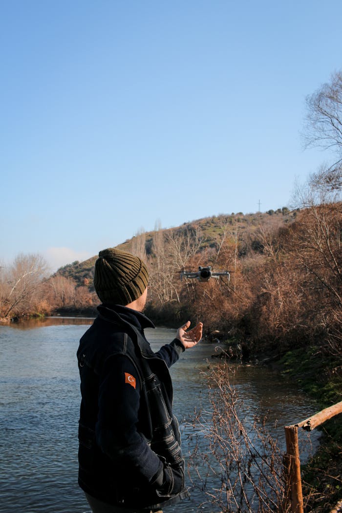 A man in a coat and hat operates a drone near a serene river surrounded by winter foliage.